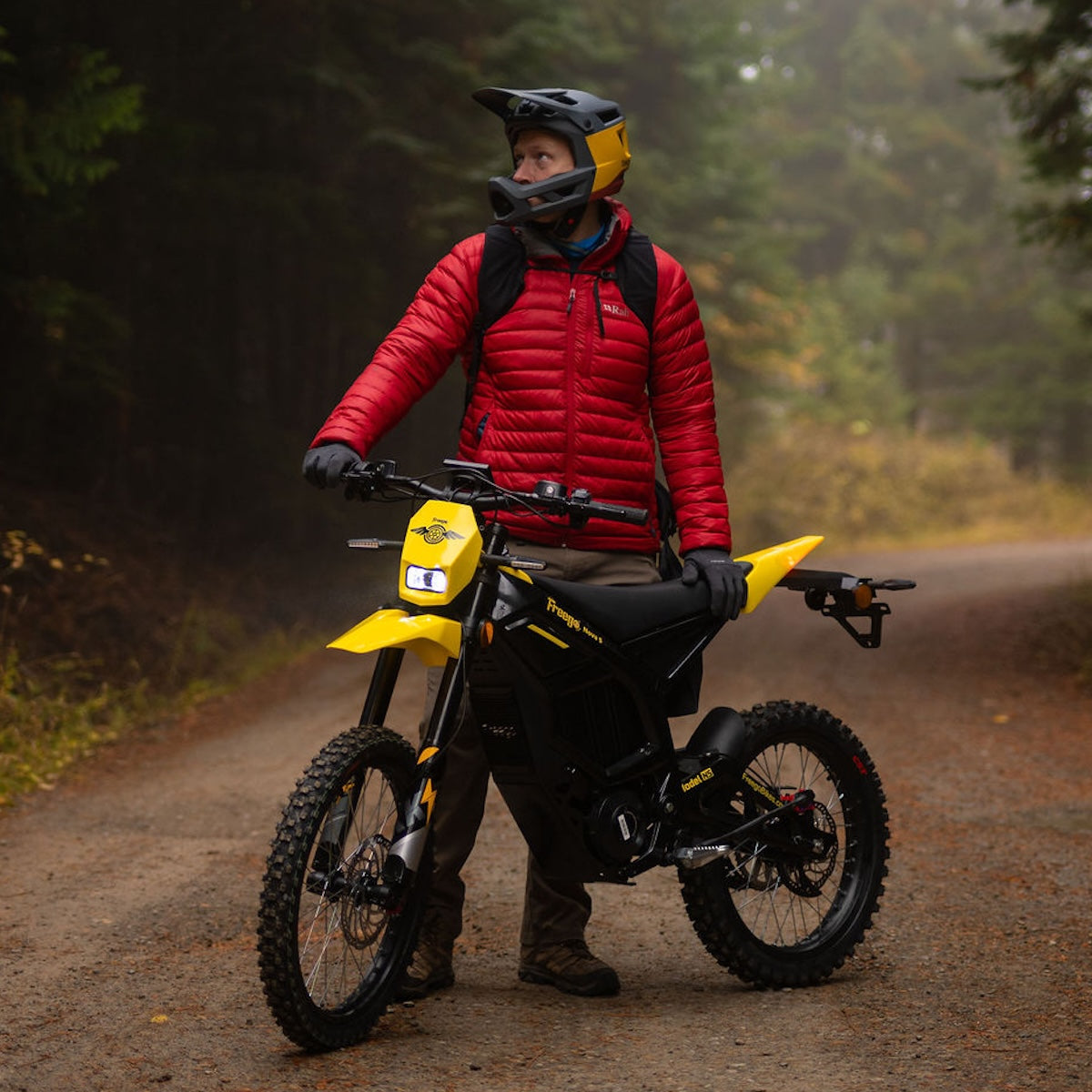 Person in a red jacket standing next to a yellow motorcycle on a forest path