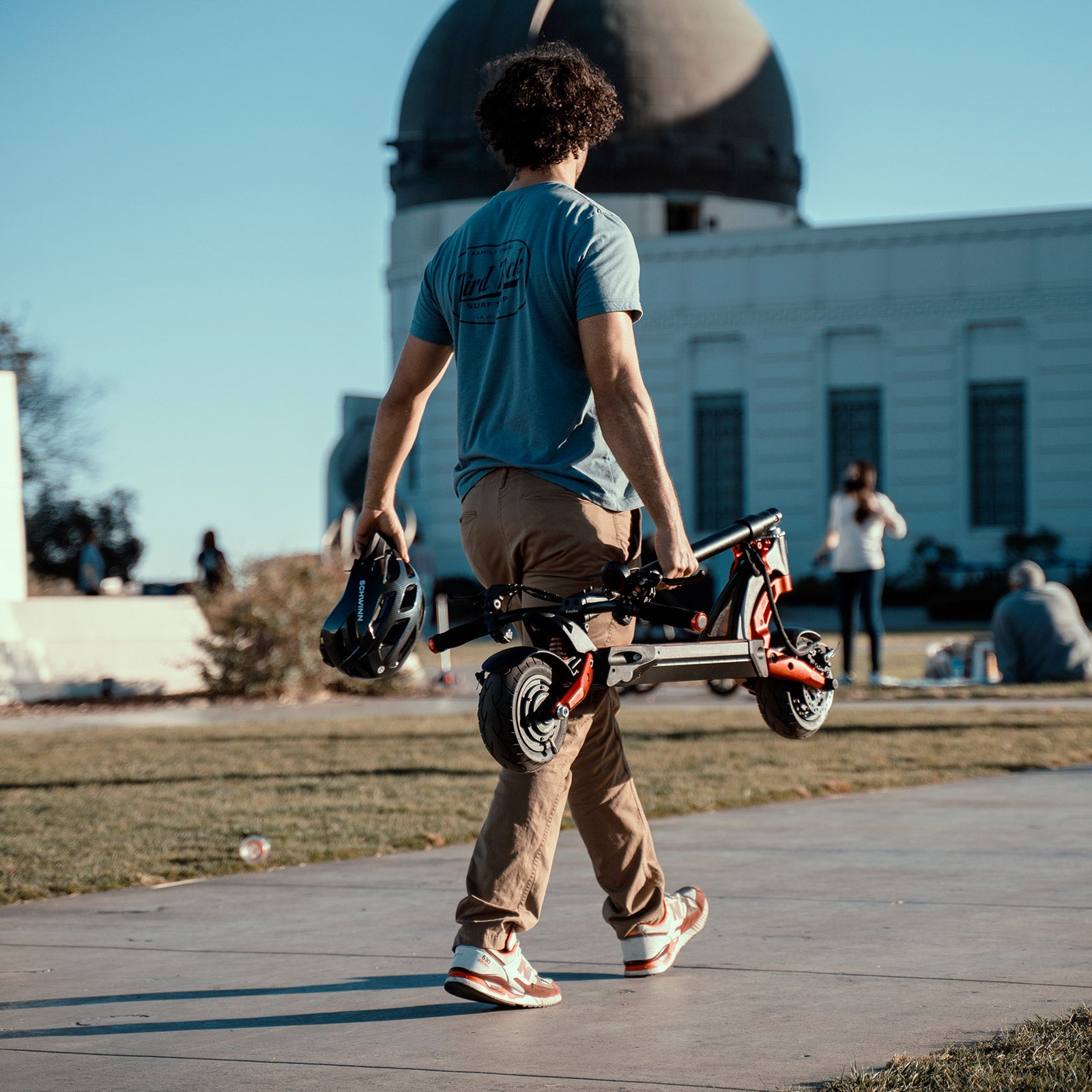 Person carrying folded Kaabo Mantis 8 Lite electric scooter in one hand while holding a helmet, demonstrating its lightweight portability.