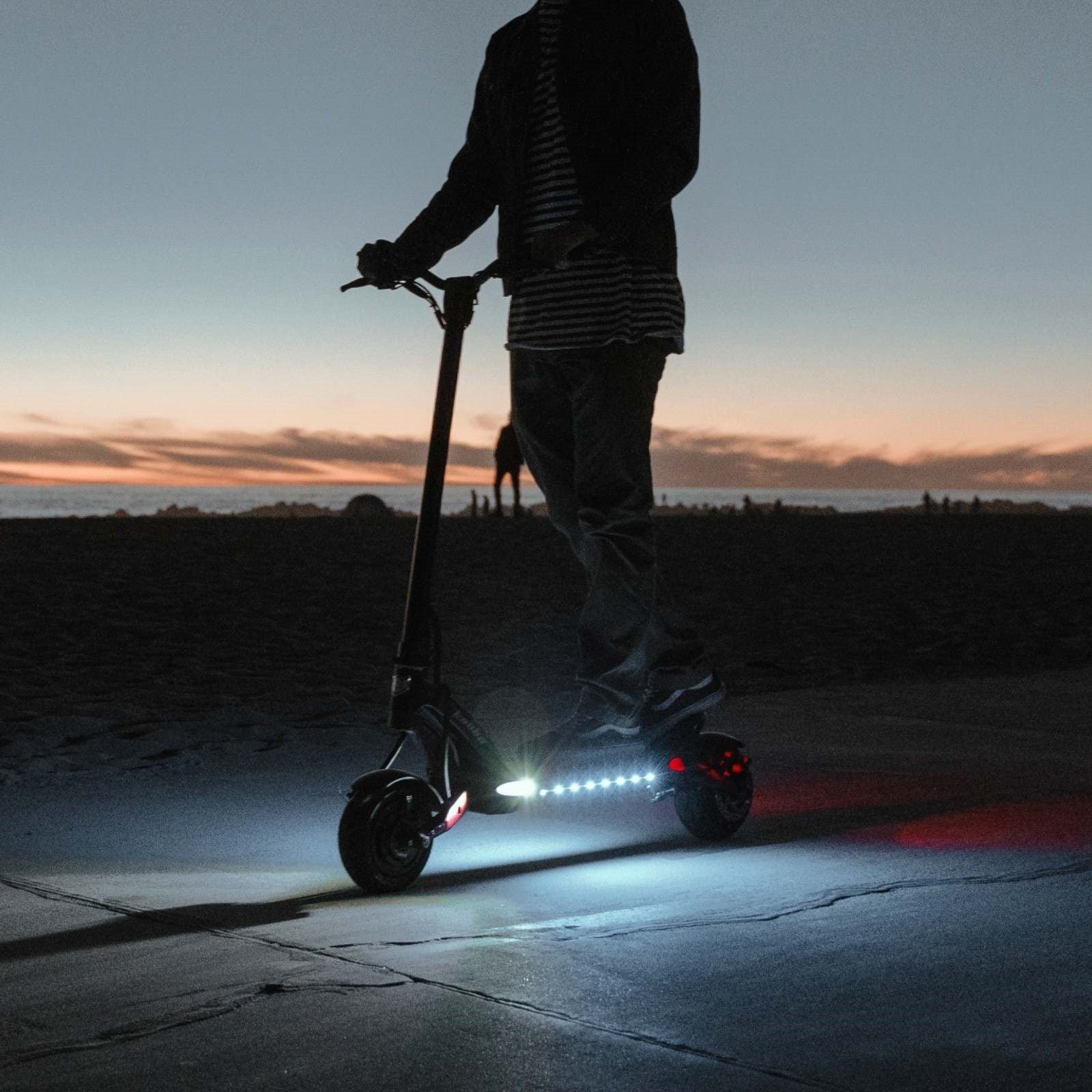 Rider using Kaabo Mantis 8 Lite electric scooter at sunset with under-deck LED lights illuminating the ground.